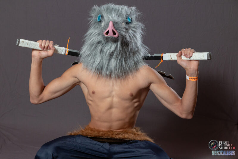 A cosplayer dressed as the boar headed Inosuke from the anime Demon Slayer crouches low with crossed swords behind his head.
