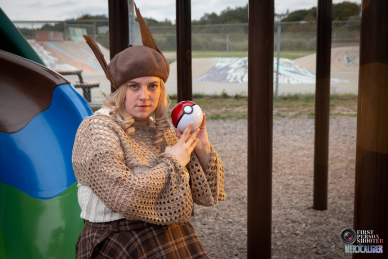 A cosplayer dressed as a schoolgirl gijinka version of the Pokemon Evee sits on a playground slide, holding a Pokeball.